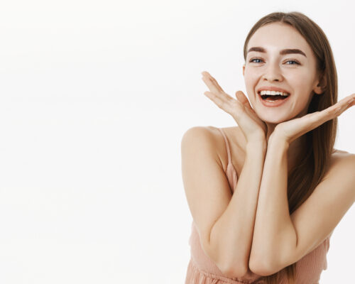 Waist-up shot of beautiful and healthy charming woman with natural beauty holding palms near jawline and smiling broadly pleased with perfect skin condition, posing cheerfully over gray background. Skincare concept